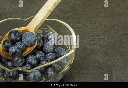 Bol en verre avec des fruits les bleuets et vieille cuillère en bois sur la pierre, sombre tableau. Cloes vue horizontale par le haut. Banque D'Images