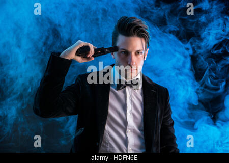 Young handsome man in suit holding gun au temple et à tout droit sur fond noir de fumée. Banque D'Images