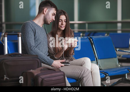 Jeune couple à l'écoute de la musique via smart phone lors de l'attente dans l'aéroport Banque D'Images