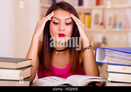 Jeune brunette woman wearing pink top séance par 24 avec pile de livres mis sur elle, tenant les mains sur la tête, fatigué l'expression du visage et de la langue, bodu concept étudiant Banque D'Images