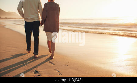 Vue arrière cropped shot of a senior couple holding hands marche sur la plage. D'âge mûr et de prendre une promenade sur le bord de la mer au coucher du soleil. Banque D'Images