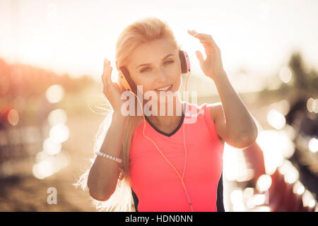Jeune femme dans le sport bénéficie d'usure et sourit à l'écoute de la musique avec des écouteurs orange. Elle met ses mains jusqu'à profiter de la vie. Banque D'Images
