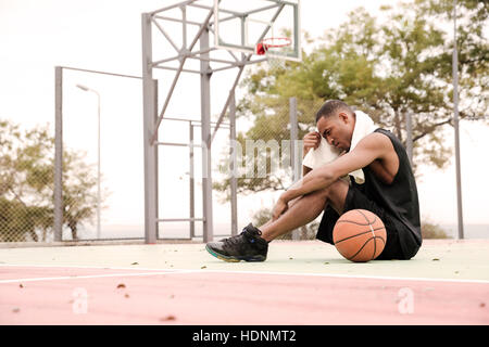 Image d'un joueur de basket-ball africain fatigué assis dans le parc avec une serviette. Banque D'Images