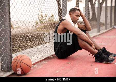 Photo de joueur de basket-ball africain fatigué assis dans le parc avec une serviette près de basket-ball. Regarder de côté. Tenant sa tête avec les mains. Banque D'Images