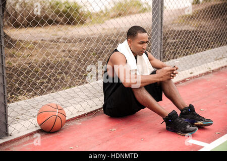 Image de jeune joueur de basket-ball africain fatigué assis dans le parc avec une serviette près de basket-ball et le chat. Regard sur téléphone. Banque D'Images