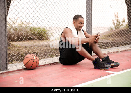 Photo de jeune joueur de basket-ball de l'Afrique de l'agréable assis dans le parc avec une serviette près de basket-ball et le chat. Regard sur téléphone. Banque D'Images