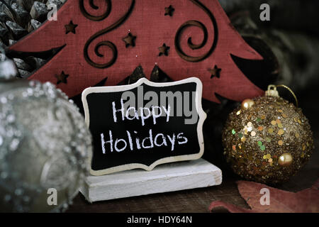 Quelques boules de noël et une pancarte noire avec le texte happy holidays écrit en C, à côté d'un arbre de Noël en bois et quelques pommes de pin Banque D'Images