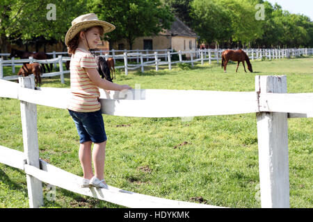 Little girl with cowboy hat standing sur corral et regarder les chevaux Banque D'Images