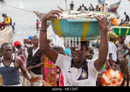 Jeunes hommes transportant de lourds bols de poisson du bateau à la rive. Les femmes attendent d'autres bateaux de pêche pour arriver. Tombo, Sierra Leone Banque D'Images