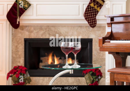 Deux verres de vin rouge sur le dessus de table en verre lumineux avec cheminée, piano et des décorations de Noël en arrière-plan Banque D'Images