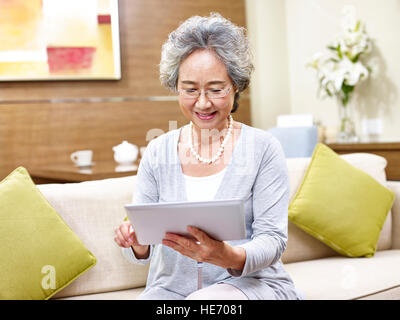 Senior asian woman sitting on sofa at home à l'aide d'un ordinateur tablette. Banque D'Images