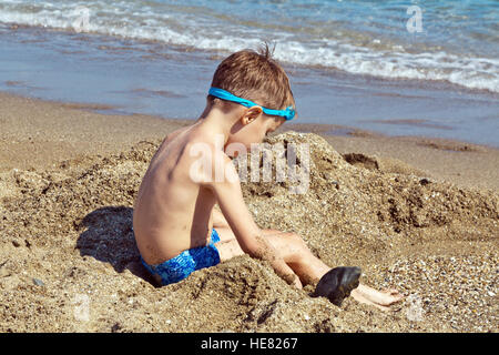Un garçon dans un maillot bleu et bleu verres pour la natation, c'est assis sur le sable à la plage
