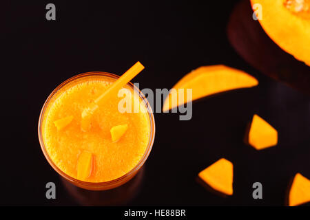 Smoothie à la citrouille dans le verre avec paille et des morceaux de feuille acrylique noire sur la citrouille avec réflexion sur fond noir Banque D'Images