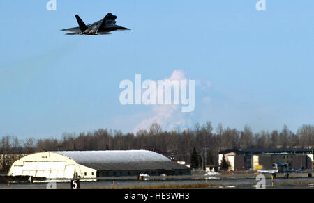 Un F-22 Raptor du 525th Fighter Squadron décolle de la base aérienne d'Elmendorf, en Alaska, près du mont Redoute, qui présente régulièrement des éruptions de vapeur. L'avion soutient les opérations dans l'Arctique et l'état de préparation du 477e groupe de chasseurs. Banque D'Images
