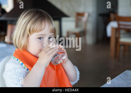 Portrait de deux ans blonde caucasien enfant avec orange bib de l'eau potable dans le verre cristal au restaurant Banque D'Images