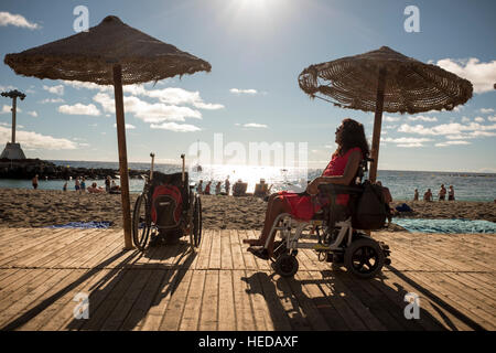 Femme dans un fauteuil roulant électrique photographié sur une terrasse en bois sur le côté à regarder une plage baignée de soleil avec les gens Banque D'Images