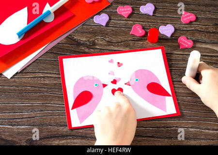 L'enfant colle les détails appliqué. Carte de voeux bébé pour la Saint Valentin. Feuilles de papier, colle, ciseaux sur une table en bois. Projet d'art pour enfants Banque D'Images