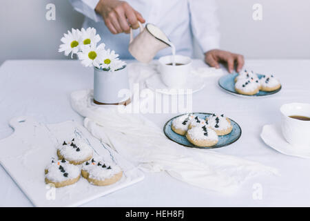 Une femme versant du lait dans une tasse de thé sur une table avec les bonshommes de cookies servis sur deux plaques et fleurs blanches dans un vase. Banque D'Images