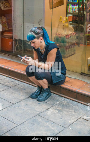 Une jeune femme , avec les cheveux teints en bleu, est assis en face de son lieu de travail en faisant une pause, fume et lit son smartphone. Banque D'Images