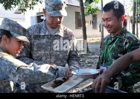 Un soldat de l'armée indonésienne et les membres de la communauté locale, offre Pvt. Marc Trinidad, un spécialiste de l'alimentaire avec le 2e Bataillon, 1e Régiment d'infanterie, 2e Stryker Brigade Combat Team, et la FPC. Voir Christina, un spécialiste de la restauration à partir de la 325e bataillon de l'aviation d'appui général de cavalerie sur Wheeler Army Airfield, Hawaii, un plat de poisson qu'ils cuits plus traditionnellement des branches de palmier au cours de l'exercice 2014 Garuda Shield. Le Garuda Shield terrain est un exercice militaire tactique bilatéral, parrainé par l'armée américaine et du Commandement du Pacifique organisée par les forces armées indonésiennes Banque D'Images