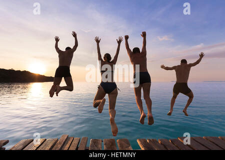Les personnes gaies s'amusant de sauter dans l'eau de mer à partir d'un quai au coucher du soleil