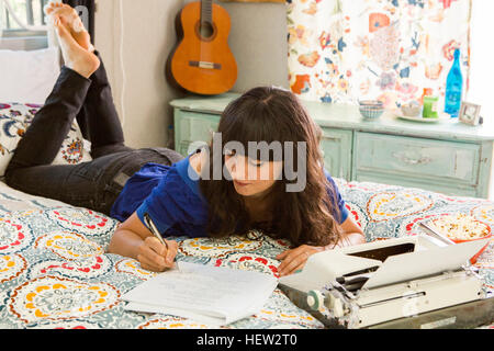 Young woman lying on bed, à l'aide de machine à écrire, à écrire des notes Banque D'Images