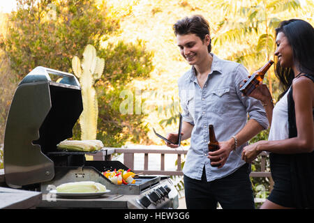 Young couple outdoors, holding de bouteilles de bière, la cuisson des aliments sur un barbecue Banque D'Images