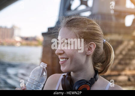 Jeune femme au bord de la rivière, portant des écouteurs autour du cou, tenant une bouteille d'eau, New York City, USA Banque D'Images