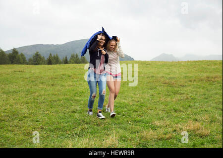 Deux jeunes femmes en marche en bas de la colline holding up anorak dans la pluie, Sattelbergalm, Tyrol, Autriche Banque D'Images