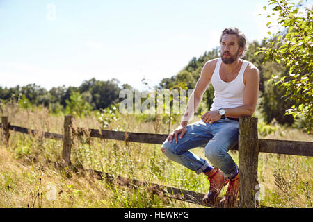 Mid adult man sitting on fence rural à out Banque D'Images