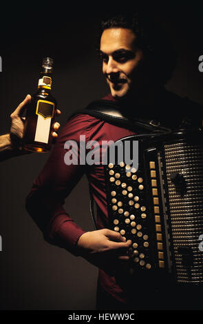 Joueur d'accordéon moderne posant avec son harmonica chromatique instrument type et regarder une bouteille d'une boisson spiritueuse dans une main sur le côté gauche. Banque D'Images