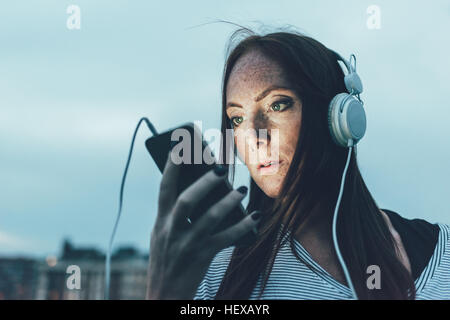 Jeune femme de rousseur listening to headphones looking at smartphone au crépuscule Banque D'Images