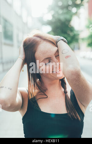 Jeune femme aux longs cheveux roux et les taches de rousseur, les yeux fermés on city street Banque D'Images