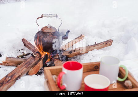 Pique-nique d'hiver romantique. Deux tasses et un bol sur un plateau en bois dans la neige. Électrique en cuivre sur un feu ouvert sur l'arrière-plan, floue. L'ébullition électrique sur firewo Banque D'Images