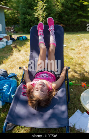 Une mignonne petite fille de sept ans met à l'envers sur une chaise bleue dans un camp à New York, United States. Banque D'Images