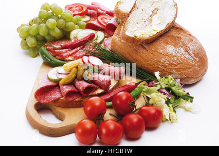 Snack-midi sur une planche à découper : salami sandwiches avec piments, cornichons, concombres et radis, garni de tomates Banque D'Images