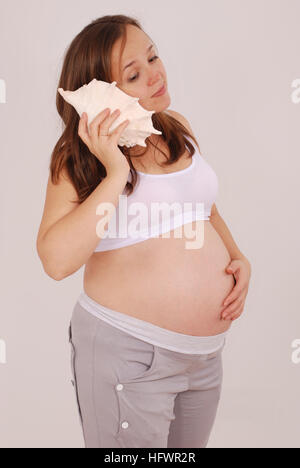 Femme enceinte a mis un coquillage à l'oreille sur fond gris Banque D'Images