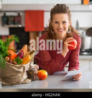 Portrait of happy young housewife holding grocery shopping vérifie dans la cuisine Banque D'Images