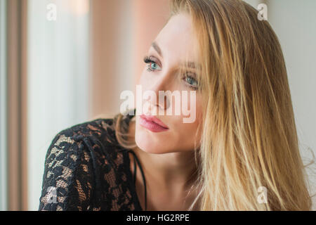 Close up portrait of beautiful young blonde woman in black lace shirt assis à côté de fenêtre, à la recherche d'émotion et réfléchi Banque D'Images