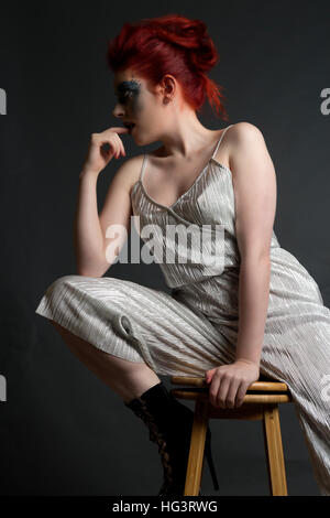 Red haired model with blue maquillage créatif portant robe d'argent, assis sur le tabouret en bois Banque D'Images