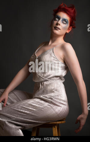 Red haired model with blue maquillage créatif portant robe d'argent, assis sur le tabouret en bois Banque D'Images