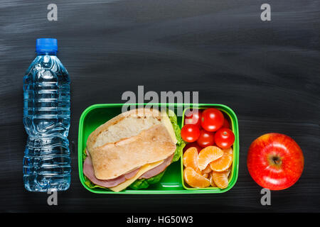 Sandwich, petites tomates, la mandarine, l'apple en plastique boîte à lunch et une bouteille d'eau sur le tableau noir. Banque D'Images