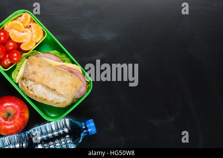 Sandwich, petites tomates, la mandarine, l'apple en plastique boîte à lunch et une bouteille d'eau sur le tableau noir. Banque D'Images