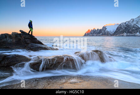 Randonneur admire les vagues de la mer de glace se brisant sur les falaises rocheuses à l'aube, Tungeneset, Senja, comté de Troms, Norvège arctique, Banque D'Images