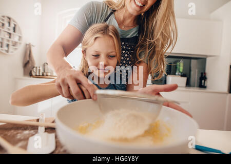 Petite fille d'apprendre à faire de la pâte à partir de sa mère. Femme en ajoutant de la farine dans un bol. Mère et fille faire de la pâte à cuire. Banque D'Images