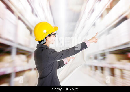 Jeune femme d'origine asiatique store manager with hard hat compte stock à l'entrepôt. Étagères avec des marchandises à l'arrière-plan. Banque D'Images
