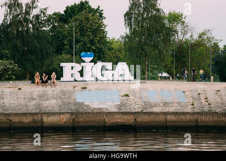 Riga, Lettonie - 30 juin 2016 : trois jeunes femmes assises sur le bord de la rivière Daugava en béton de remblai à côté de grand panneau de nom de ville. Banque D'Images