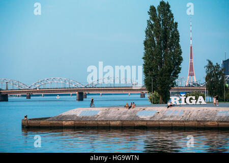 Riga, Lettonie. Le remblai à béton Daugava avec nom de ville signer et de repos les gens autour. Tour de télévision de Radio, Pierre et ponts ferroviaires Zone Banque D'Images