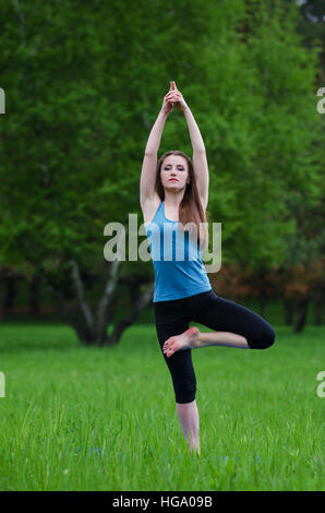 Jeune fille pratiquant le yoga dans la nature dans les bois sur un fond d'arbres et l'herbe verte. Elle équilibre sur une jambe. Banque D'Images