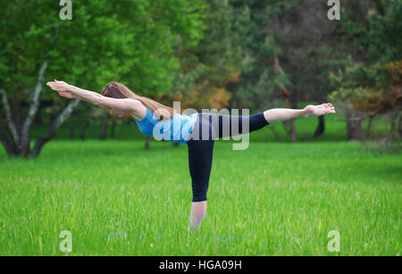 Jeune fille pratiquant le yoga dans la nature dans les bois sur un fond d'arbres et l'herbe verte. Elle équilibre sur une jambe. Banque D'Images